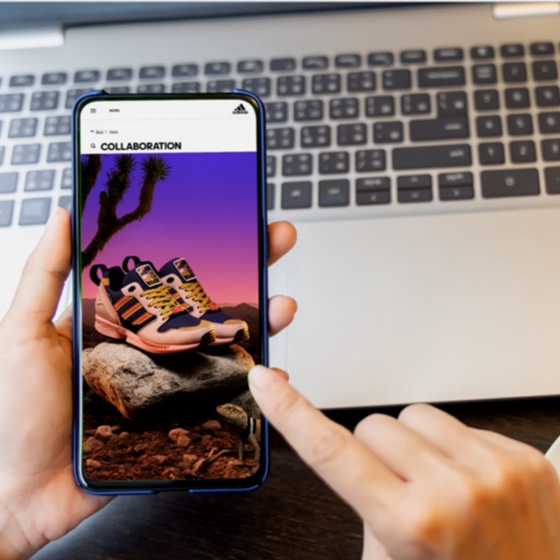 hand holding smart phone with athletic shoes on the screen in front of laptop keyboard