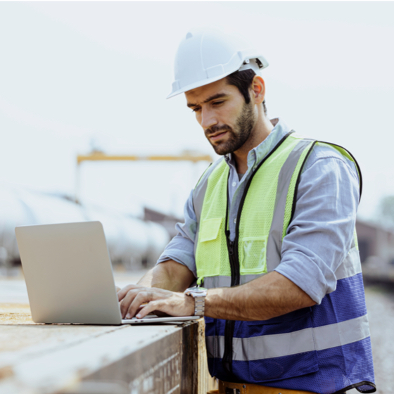 man in construction hat and vest working on a laptop at jobsite