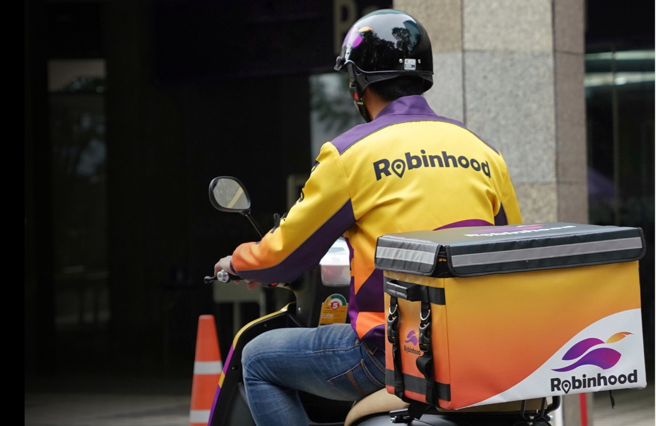 A Robinhood delivery driver on a motor scooter and wearing a Robinhood branded jacket. 