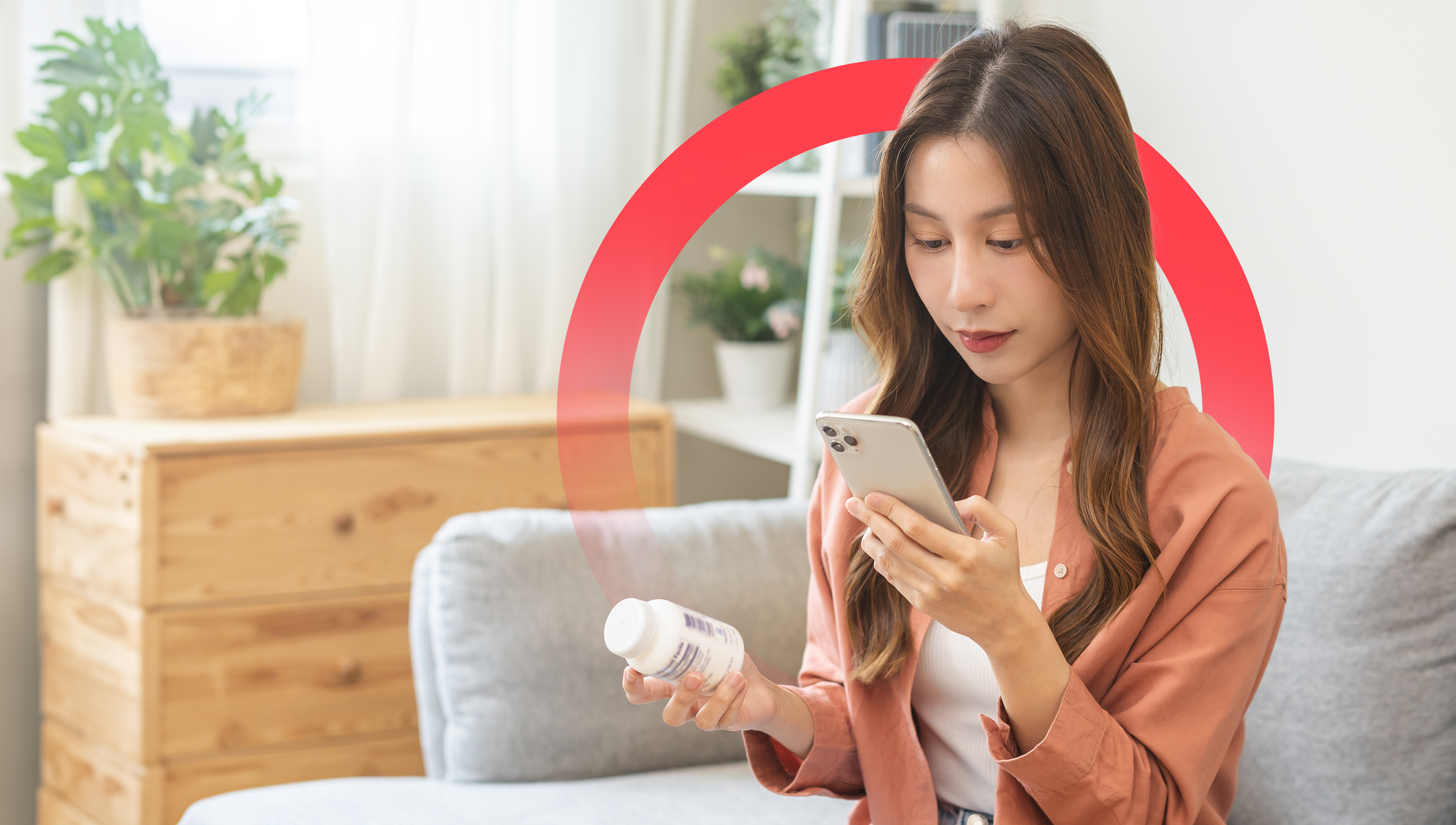 A woman sitting on her couch holds a prescription bottle in her right hand while checking her smartphone for dosage recommendations.